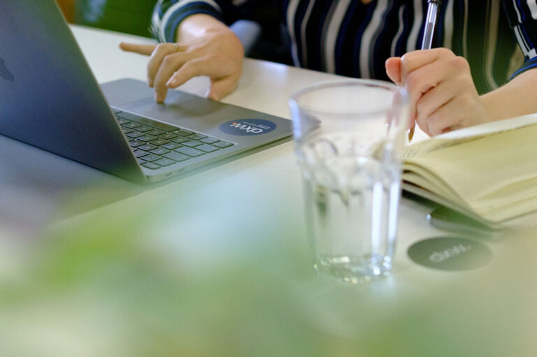 A light-skinned person in a blue striped shirt is sitting at a desk, typing on a laptop with one hand and writing in a notebook with another. There is a glass of water in front of the notebook, and blurred green foliage in the foreground.