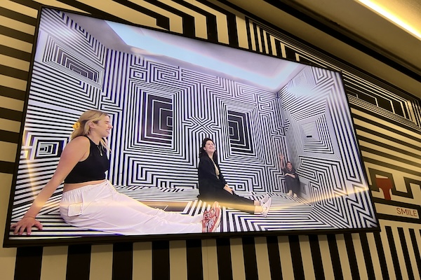 A large wall-mounted screen showing 3 members of dxw's design team in a small, brightly-lit room in the Twist Museum that has black and white lines on the walls and floor. 2 of them are sitting down with their legs outstretched and one is sitting in the corner.