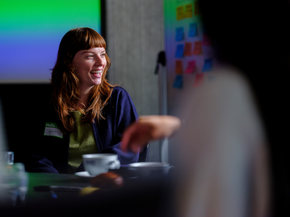 A white woman with red-brown hair wears a blue jacket. She is smiling and sitting in front of a gradient blue and green background. The foreground is blurred, showing a coffee cup on a table and another person's hand.